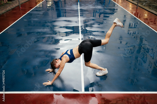 Athletic woman does sports on the street in the rain, stretching on a wet playground.