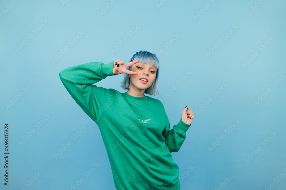 © bodnarphoto - Beautiful girl in a green sweatshirt and with colored hair poses for the camera on a blue background, shows a gesture of peace and laughs