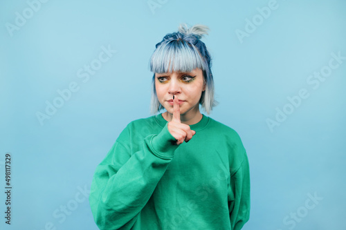 Cute teen girl with blue hair stands on a blue background, looks away and shows a gesture of silence.