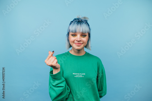 Positive hipster girl with colored hair stands on a blue background, shows his fingers a heart gesture and looks at the camera with a smile