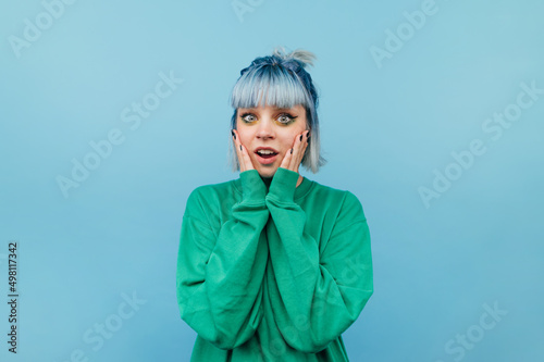 Portrait of shocked joyful hipster girl with blue hair holding her cheeks and looking at camera with happy face.