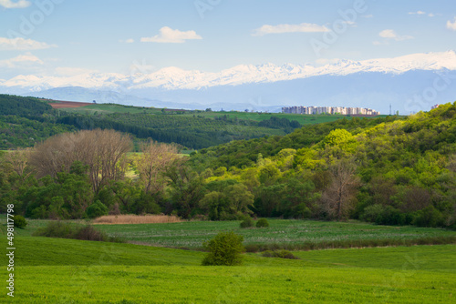 Springtime landscape, grassy field, and rolling hills. Snow-capped high mountains. A rural town in the distance. Sunny afternoon.