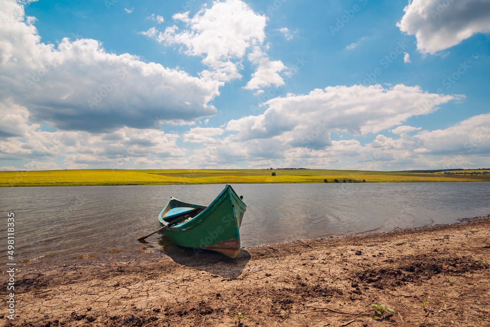 Old fishing boat on the lake shore in front of dry brown soil, behind a beautiful rural landscape with yellow fields and blue sky with clouds. Old fishing boat on the lake shore in front of dry brown soil, behind a beautiful rural landscape with yellow fields and blue sky with clouds.