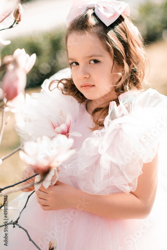 Beautiful calm kid girl 4-5 year old wear stylish princess dress smell magnolia flower over blooming tree in garden outdoor. Spring season. Childhood. Springtime. Happy child with floral nature.