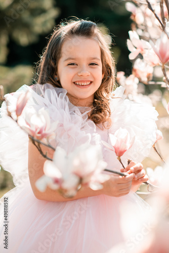 Beautiful smiling kid girl 4-5 year old wear stylish princess dress smell magnolia flower over blooming tree in garden outdoor. Spring season. Childhood. Springtime. Happy child with floral nature.