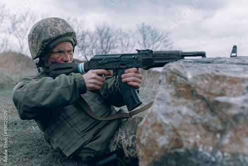 Ukrainian soldier shoots from kalashnikov assault rifle to russian enemy near rock.