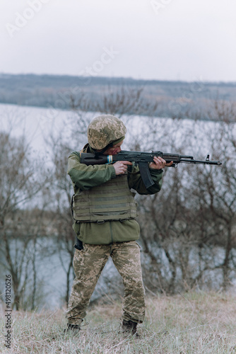 Ukrainian soldier aims from kalashnikov assault rifle to russian enemy against background of river and sky.