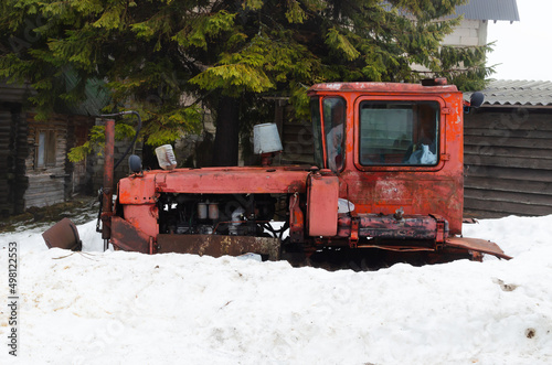 An old red small bulldozer stands in the snow among tall pines. Rural life concept. Spring in the mountains. Horizontal orientation.