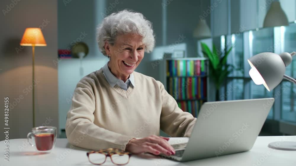 Happy elderly woman using laptop and smiling sitting in spacious living room at home