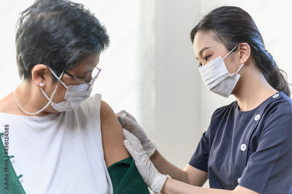 Young Asian woman nurse injecting covid-19 vaccine to senior Thai woman patient wearing mask in clinic or health care center. Coronavirus pandemic protection or health care medical concept. Young Asian woman nurse injecting covid-19 vaccine to senior Thai woman patient wearing mask in clinic or health care center. Coronavirus pandemic protection or health care medical concept.