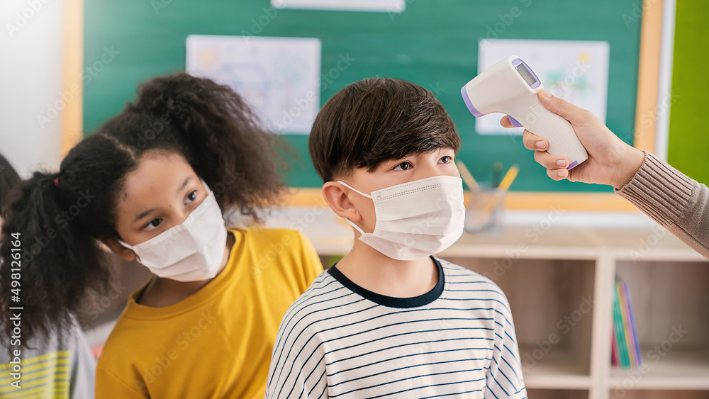 Portrait of students in medical mask in classroom, covid-19 outbreak. Teacher kids with thermometer at preschool entrance. Social distancing, coronavirus prevention. Temperature check in at school.