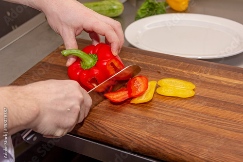 The cook cuts a sweet pepper with a knife on a wooden board