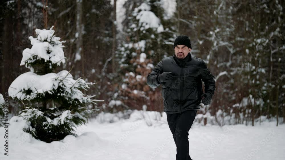 adult man in black clothes is running in snowy forest in daytime, training her body and endurance adult man in black clothes is running in snowy forest in daytime, training her body and endurance