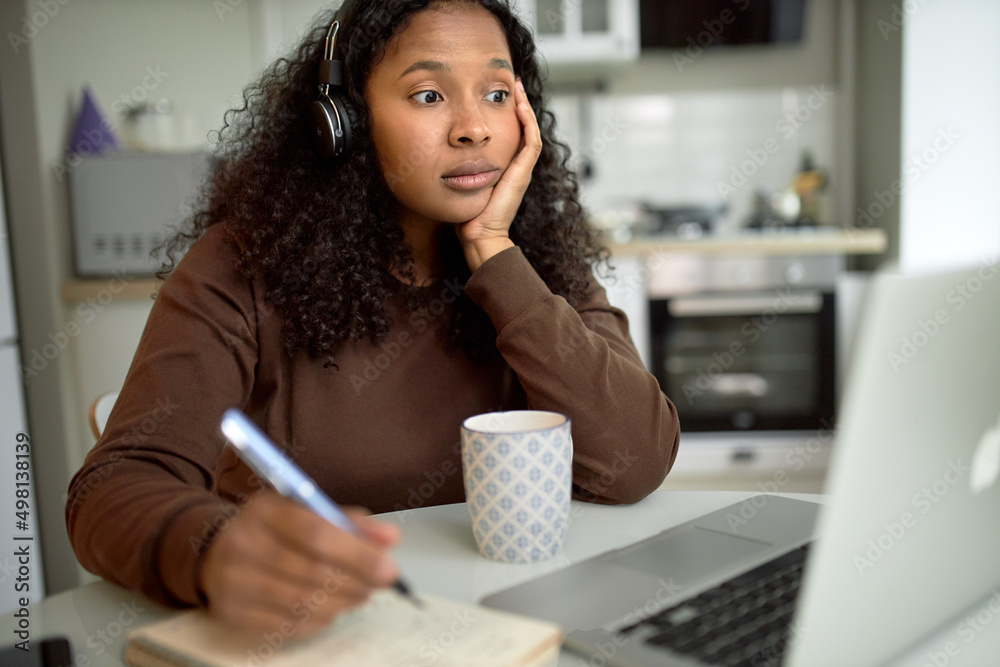 Surprised African millennial female student in headphones making notes ...