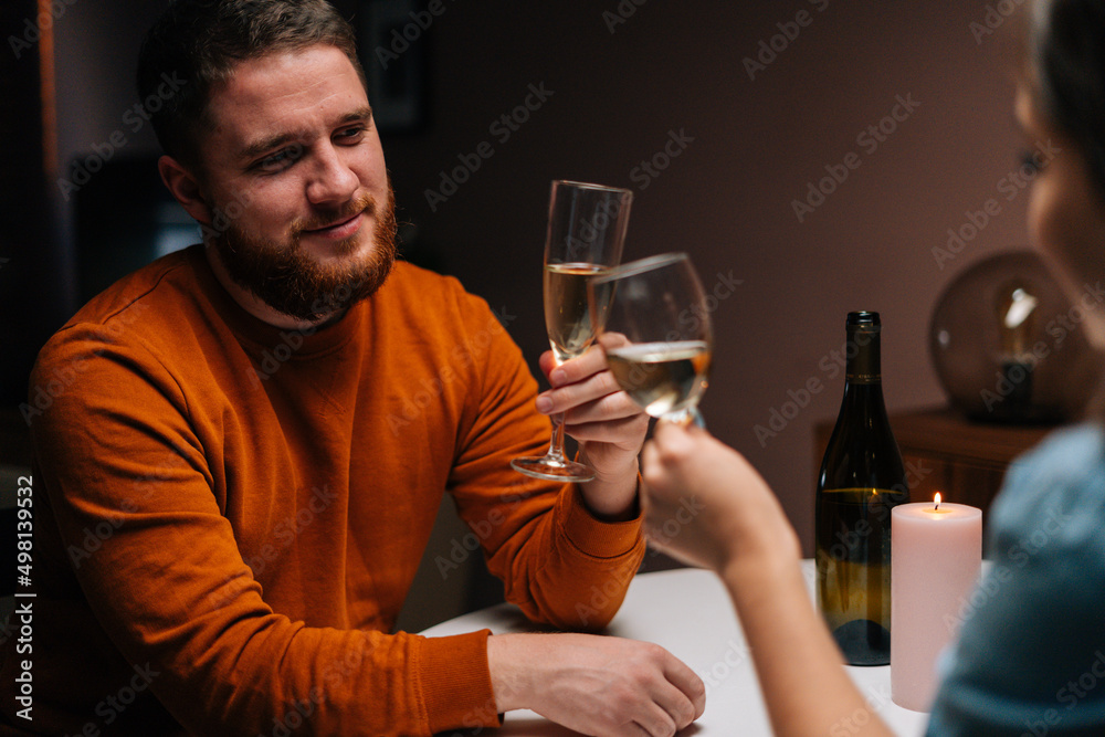 Close-up of happy young couple clinking glasses of white wine sitting at table with candles in dark cozy room. Love couple celebrating anniversary, Valentines day, enjoying romantic date.