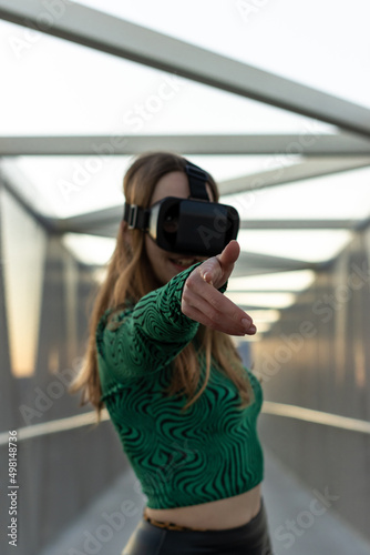 Young hispanic woman wearing in virtual reality goggles, touch something like push click on button, pointing at floating virtual screen outdoors. lifestyle concept. Vertical portrait. Selective focus
