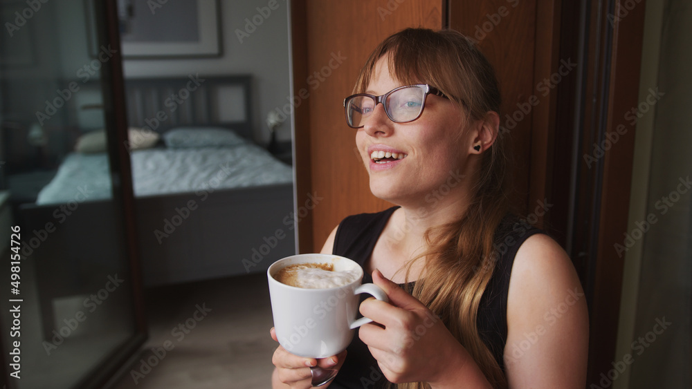 Closeup portrait of a millennial European woman holding a big cup of cappuccino coffee, looking at her neighbors, and smiling. Bedroom in the background. High quality photo Closeup portrait of a millennial European woman holding a big cup of cappuccino coffee, looking at her neighbors, and smiling. Bedroom in the background. High quality photo