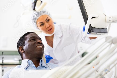 Professional female dentist showing smiling african american patient x-ray of teeth on computer screen, explaining good results after dental treatments