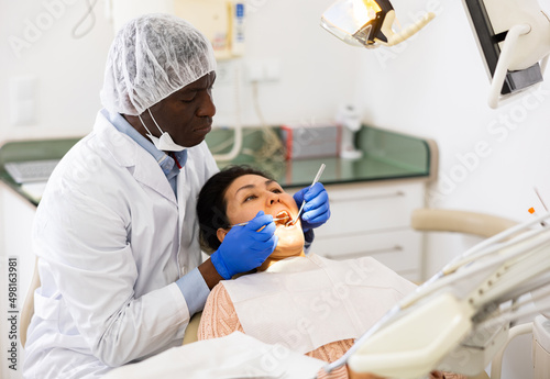 Professional african american dentist performing teeth treatment to adult woman in dental clinic..