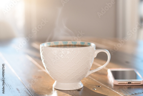 Картината върху платно White cup of hot steaming coffee on table with mobile phone and morning sunlight
