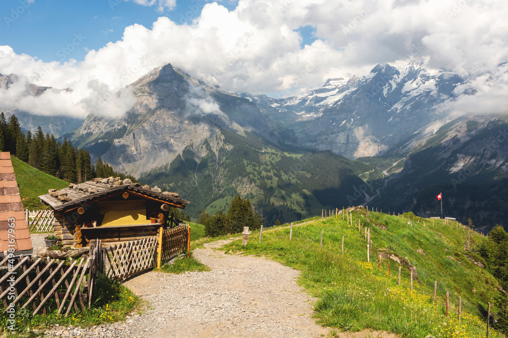 Beautiful Swiss mountains in white clouds, snow on the peaks and green meadows, a sandy stone path along a wooden house with a fence to the red national flag Beautiful Swiss mountains in white clouds, snow on the peaks and green meadows, a sandy stone path along a wooden house with a fence to the red national flag