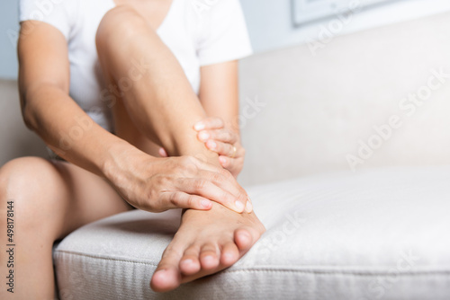 Foot pain, Asian woman sitting on sofa hold her ankle injury feeling pain in her foot at home, female suffering from feet ache use hand massage relax muscle from ankle interior, Healthcare and medical