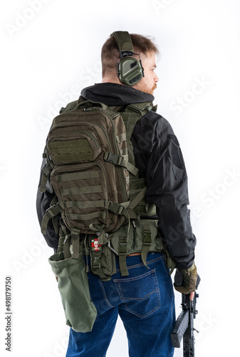 airsoft player in full growth. a man in an outfit, in headphones, a bulletproof vest, with a rifle and a pistol on a white background. standing with his back to us