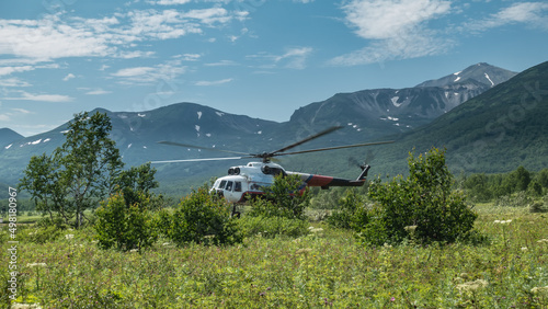 The helicopter is on the landing pad on a green alpine meadow. There is lush grass and trees around. A picturesque mountain range against the blue sky. Kamchatka. Nalychevo photo