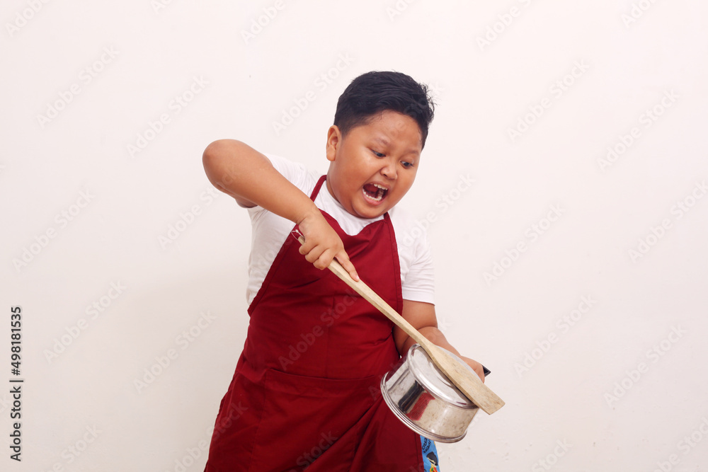 Angry asian boy with apron standing while holding a kitchen ware. Cooking class concept Angry asian boy with apron standing while holding a kitchen ware. Cooking class concept