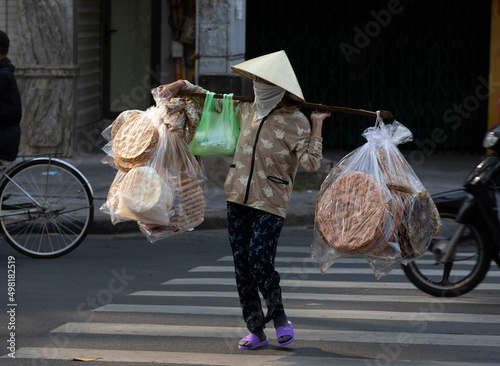 A Vietnamese woman with traditional hat selling flat bread crossing the road with her equipment and stall on her back