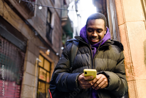 Low angle of cheerful black man in casual outerwear with backpack. He is smiling and reading text messages on cellphone while leaning on wall on blurred background of city street