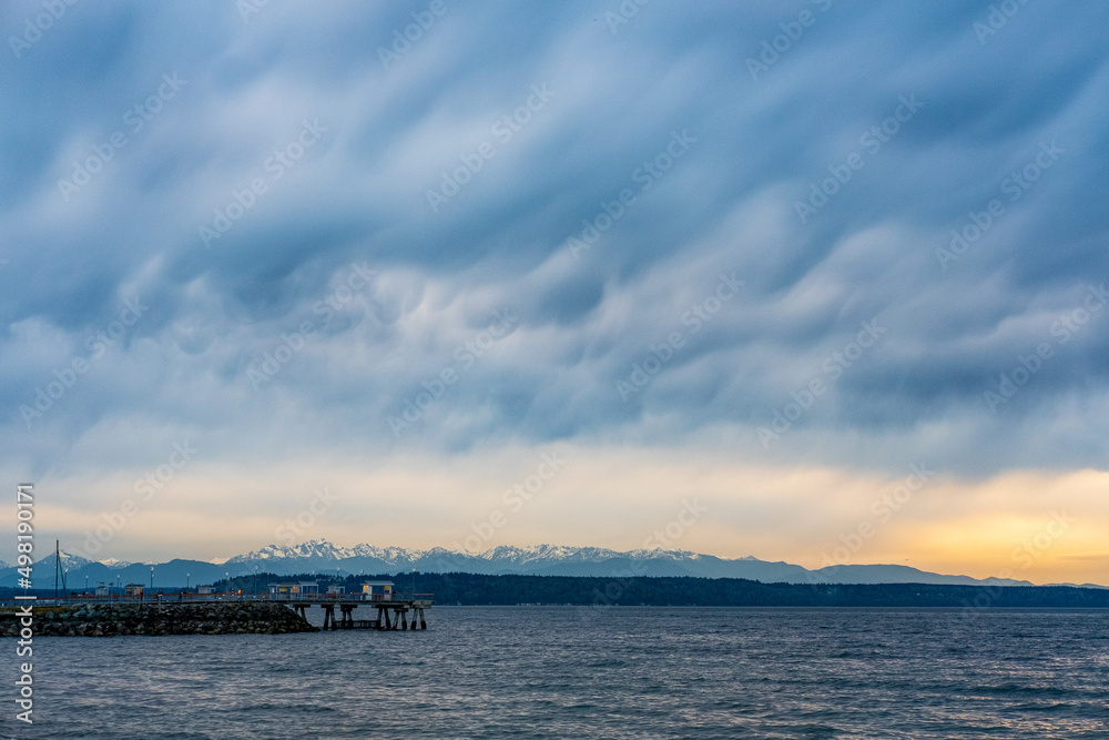 Complex clouds and stark mountains overshadow a fishing pier on Puget Sound