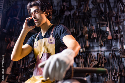 Принти на полотні Authentic blacksmith man wear leather apron in dark indoors studio calling by ph