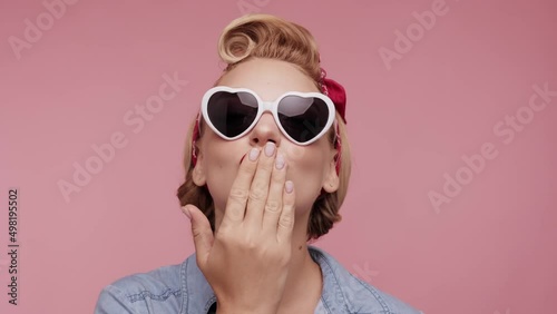Pretty Pin-up blond Girl Look at the camera, lowers her glasses, blinking and sending air kiss. Pretty Charming woman in denim jacket posing isolated on pink color background. Retro female.