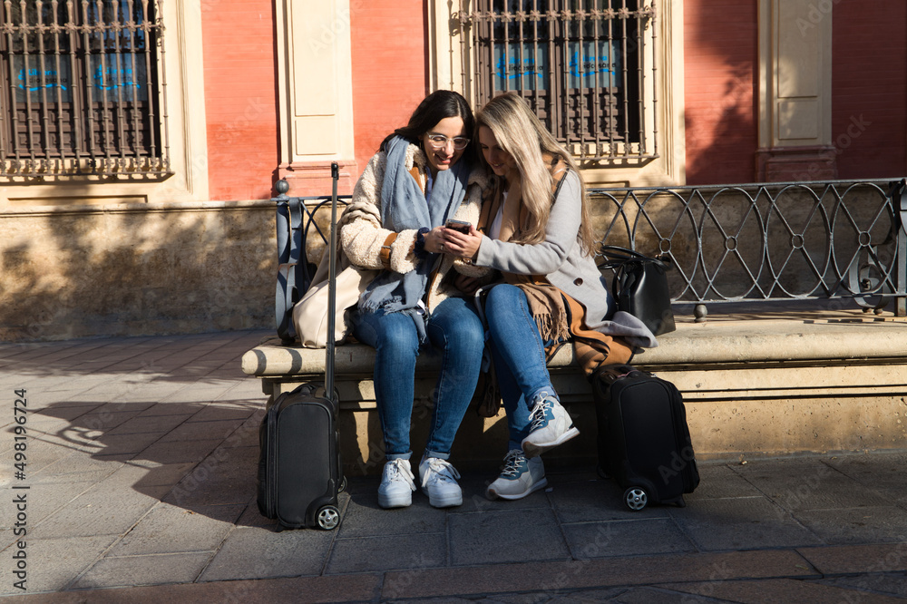 lesbian couple sitting on a bench in a monumental square in the city. They are consulting their mobile phones when they arrive in the city on holiday and their suitcases are next to them. Tourism.
