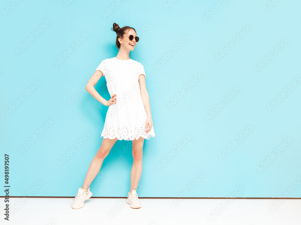 Portrait of young beautiful smiling female in trendy summer white dress. carefree woman posing near blue wall in studio. Positive model having fun indoors. Cheerful and happy. Full length Portrait of young beautiful smiling female in trendy summer white dress. carefree woman posing near blue wall in studio. Positive model having fun indoors. Cheerful and happy. Full length