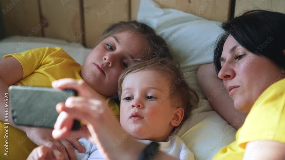 Happy family concept. Mom, daughter and son are play on bed. Child watching video on smartphone. Family leisure at home. Boy and girl with mom are watching movie on smartphone. Home education concept Happy family concept. Mom, daughter and son are play on bed. Child watching video on smartphone. Family leisure at home. Boy and girl with mom are watching movie on smartphone. Home education concept