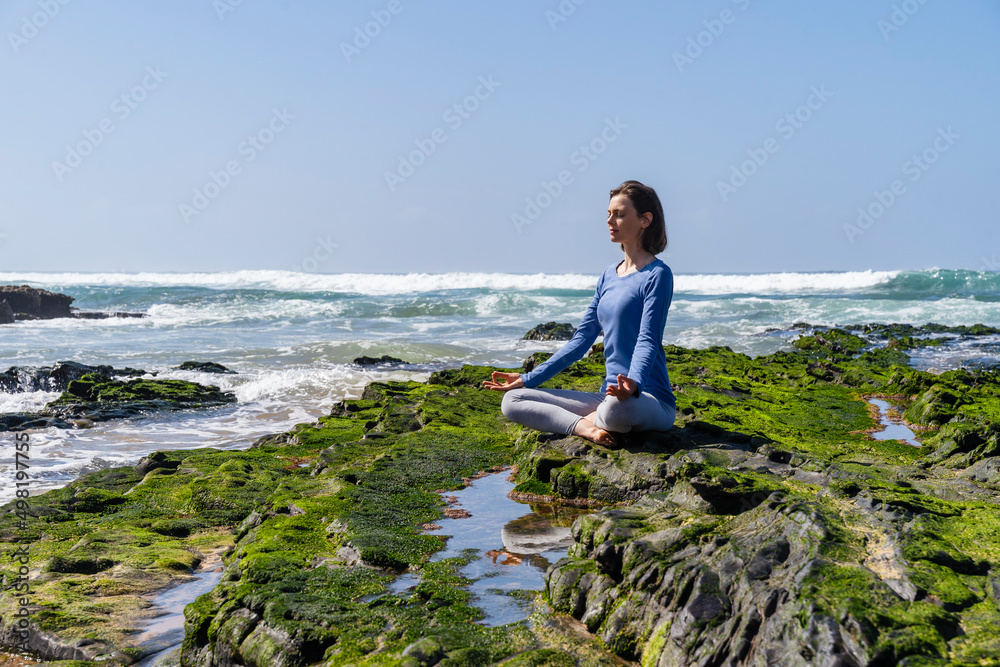 Woman sitting in lotus position and meditating on rock at beach Woman sitting in lotus position and meditating on rock at beach