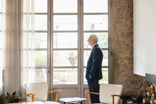 Man with hand in pocket looking through widow standing in living room at home