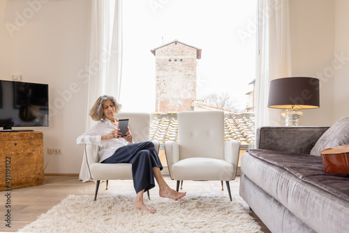 Senior woman with tablet PC sitting on armchair in living room at home photo