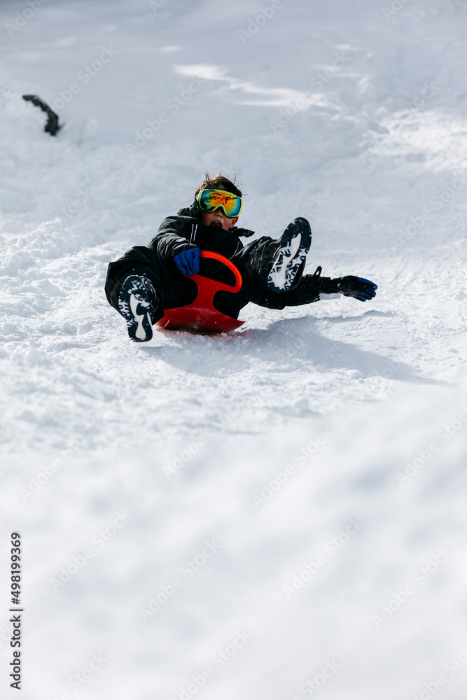 Playful boy wearing ski goggles tobogganing through snow Playful boy wearing ski goggles tobogganing through snow