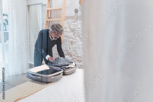 Senior freelancer packing suitcase in bedroom seen through glass photo