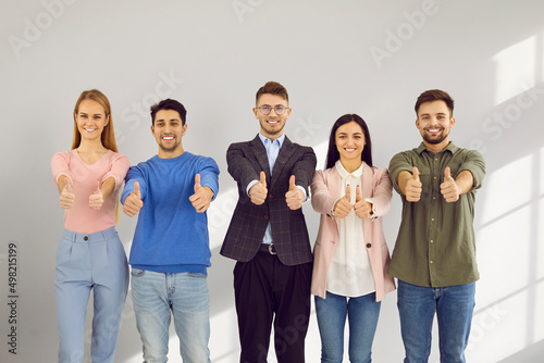 Portrait of active and smart young people showing thumbs up approving or recommending. Smiling diverse men and women casual clothes stand in row on gray wall background. Concept of positive approval