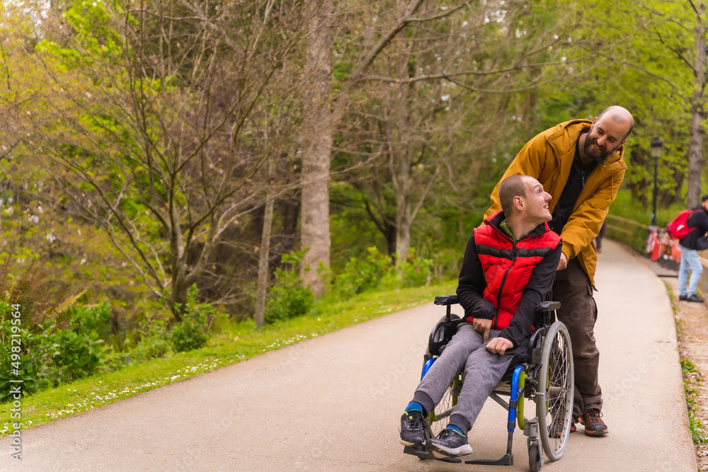 Naklejka premium Paralyzed young man in the wheelchair being pushed by a friend in a public city park, strolling along a path having fun in spring