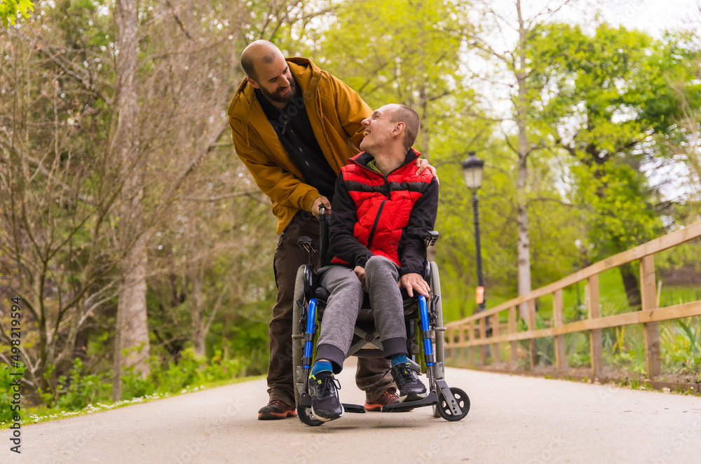 Paralyzed young man in the wheelchair being pushed by a friend in a public city park, strolling along a path having fun and smiling Paralyzed young man in the wheelchair being pushed by a friend in a public city park, strolling along a path having fun and smiling