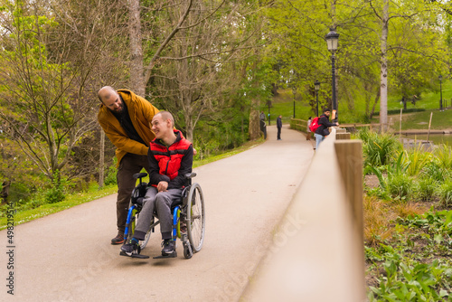 Paralyzed young man in a wheelchair being pushed by a friend in a public city park, strolling along a path