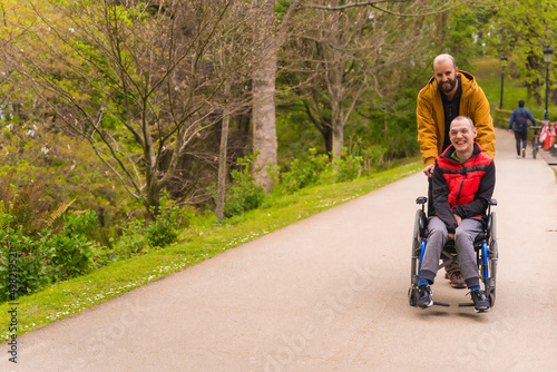 Paralyzed young man in a wheelchair being pushed by a friend in a public city park, strolling along a path © unai