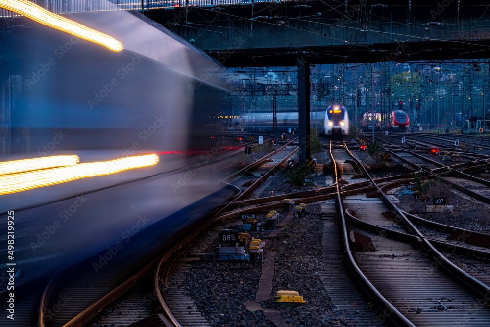 Naklejka premium Blue hour at the main station of Hagen Germany with many lights, signals, catenary, glistening tracks, switches and trains in motion. Railway infrastructure and technology at morning twilight. 