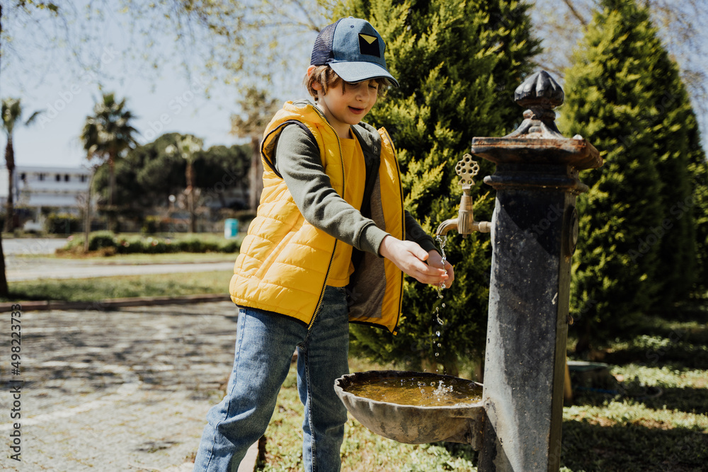 Kid schoolboy in casual clothing washing his hands in the street old fashioned drinking water fountain. Boy in blue jeans and yellow vest playing with water from drinking sprinkler in city park.
