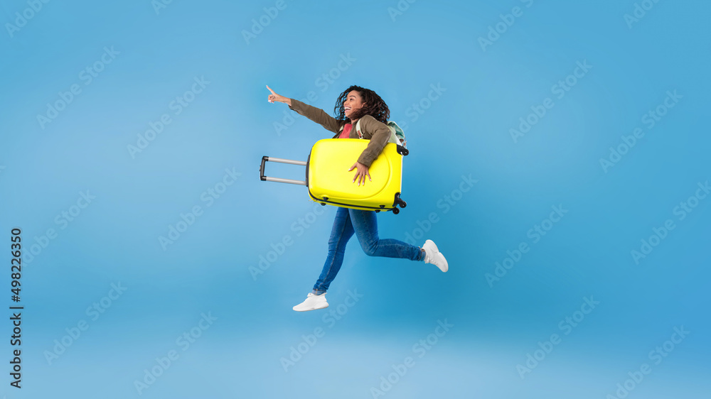Happy young black woman jumping with suitcase and pointing at blank space for travel agency ad on blue background